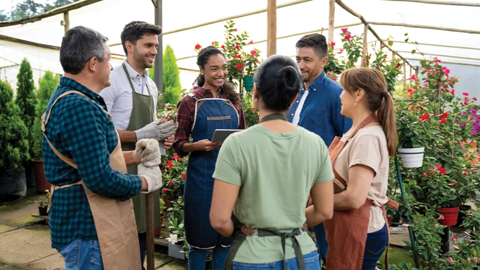 A group of six people gather in a circle standing in a greenhouse surrounded by flowers.
