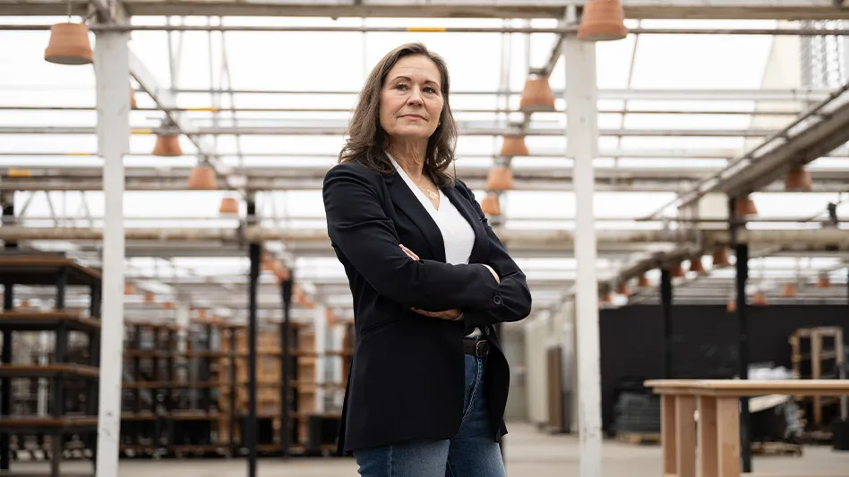 A woman wearing a black suit jacket, white T-shirt and blue jeans stands with her arms crossed and looking off-camera. In the background is a greenhouse with empty shelves and pots.