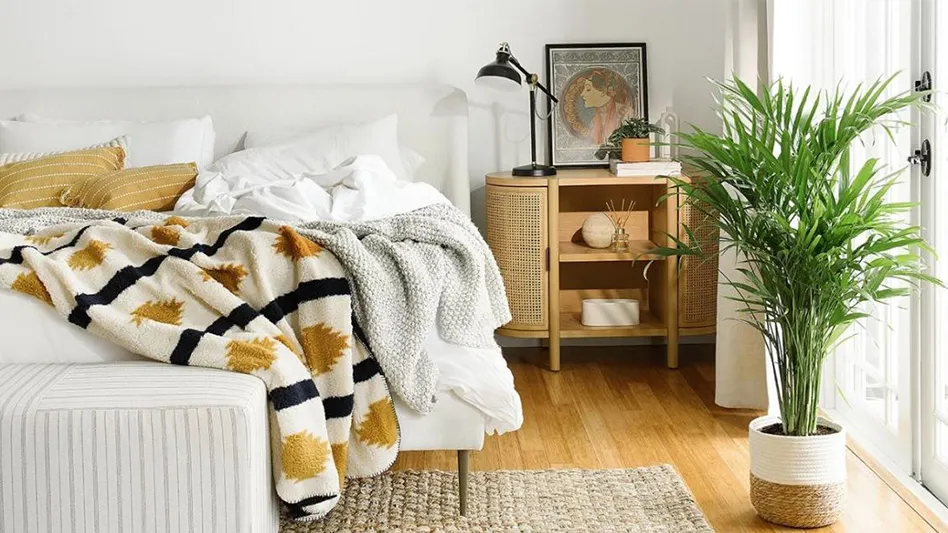 A tall green plant in a white and tan rope pot sits in a bedroom. There's a white bed with black, tan, cream and gray bedding, a gray and white bench at the foot of the bed, a tan rug on the orange hardwood floors, a brown wooden end table with a black lamp and a white patio door.