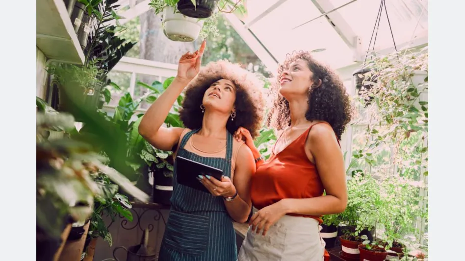 A garden center employee carrying an iPad points to a plant to show a customer.