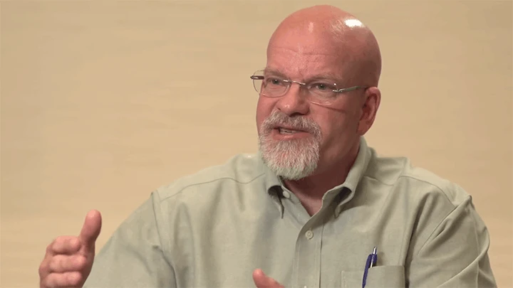 A bald man with a white beard wearing glasses and a tan button-up shirt gestures with both hands as he talks.