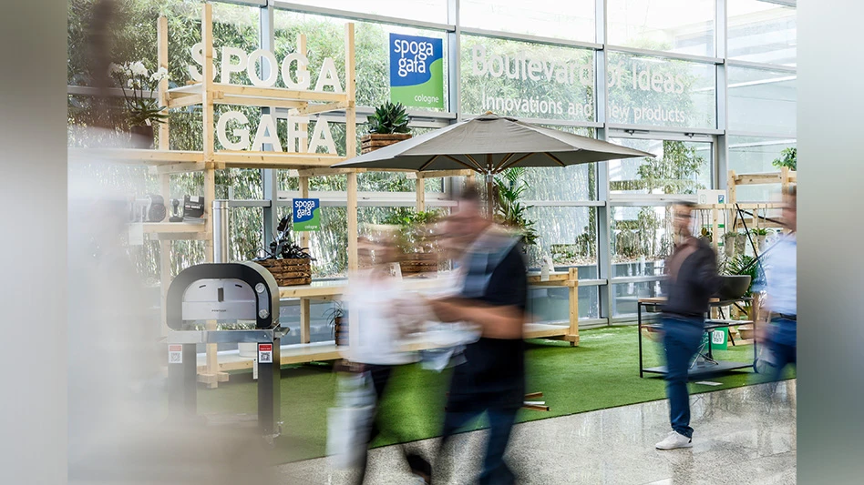 Blurred people walk past multiple signs that say spoga+gafa Cologne, as well as a sign that says Boulevard of Ideas Innovation and New Products, with products including a grill, an umbrella, tables and plants sitting on artificial grass.