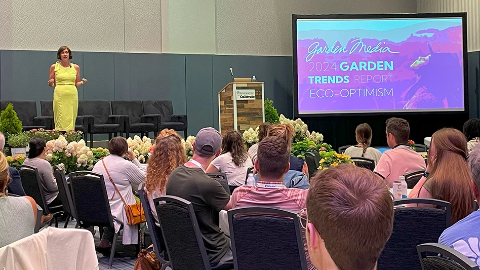 A woman wearing a lime green dress stands on a stage in front of a group of people sitting in black chairs at tables covered in white tablecloths. On the stage to the right of the woman is a brown podium with a sign that says AmericanHort Cultivate, and to the right of the podium is a large presentation screen that says Garden Media 2023 Garden Trends Report Eco-optimism.