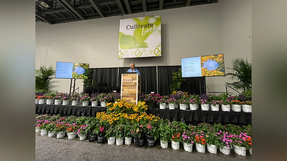 A man stands on a stage surrounded by many containers of plants. Above him is a sign that says Cultivate Live!