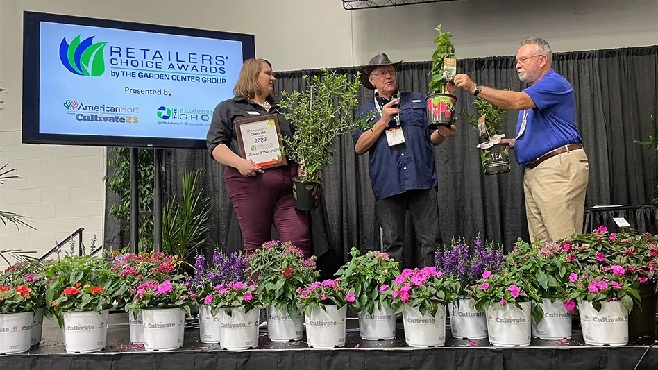 Three people stand on a stage with multiple containers of flowers in front of them.