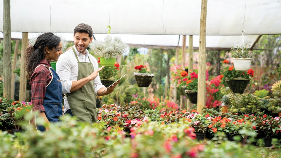 A smiling man and a woman walk through a nursery looking at a clipboard in the man's hand.