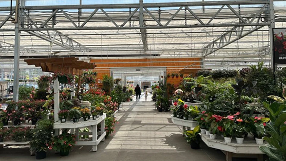 A person and child walk through the doors of the outdoor area of a garden center that's full of green plants on wooden shelves.