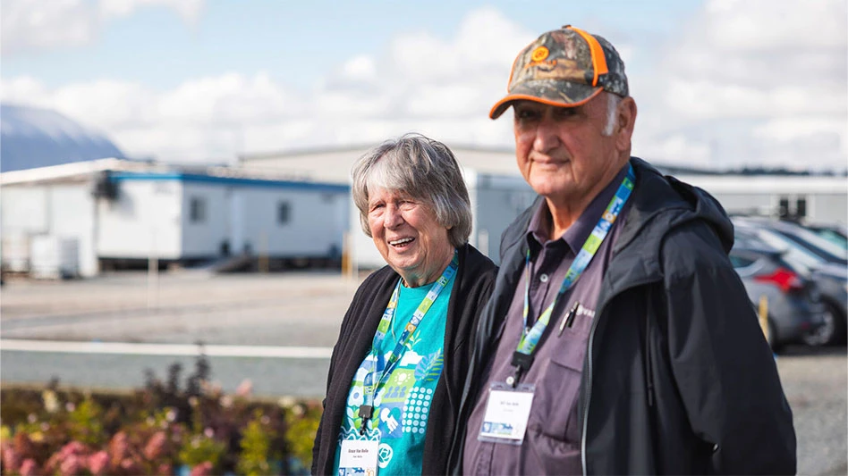 A woman with short grey hair and a man with a baseball cap on stand in a parking lot in front of warehouses.