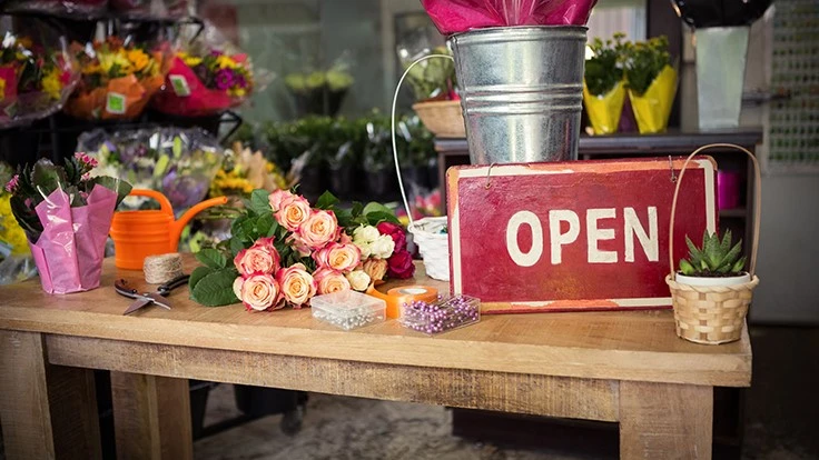 Flowers and a red open sign sit on a wooden table.